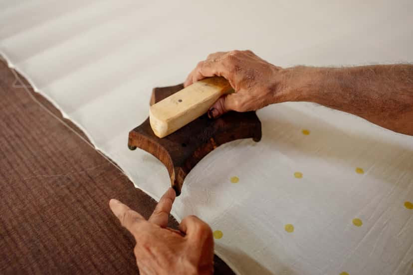 An artisan's hands pressing a carved wooden block onto ivory cloth